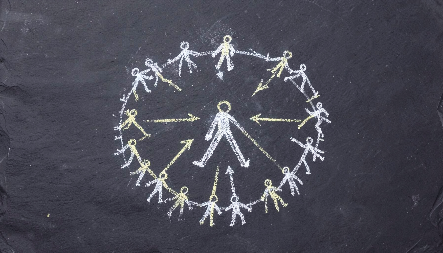 A chalk drawing on a dark slate board showing a circle of stick figures connected by arrows, with a larger central figure guiding the group, drawn in white and pale yellow chalk against the deep charcoal background.