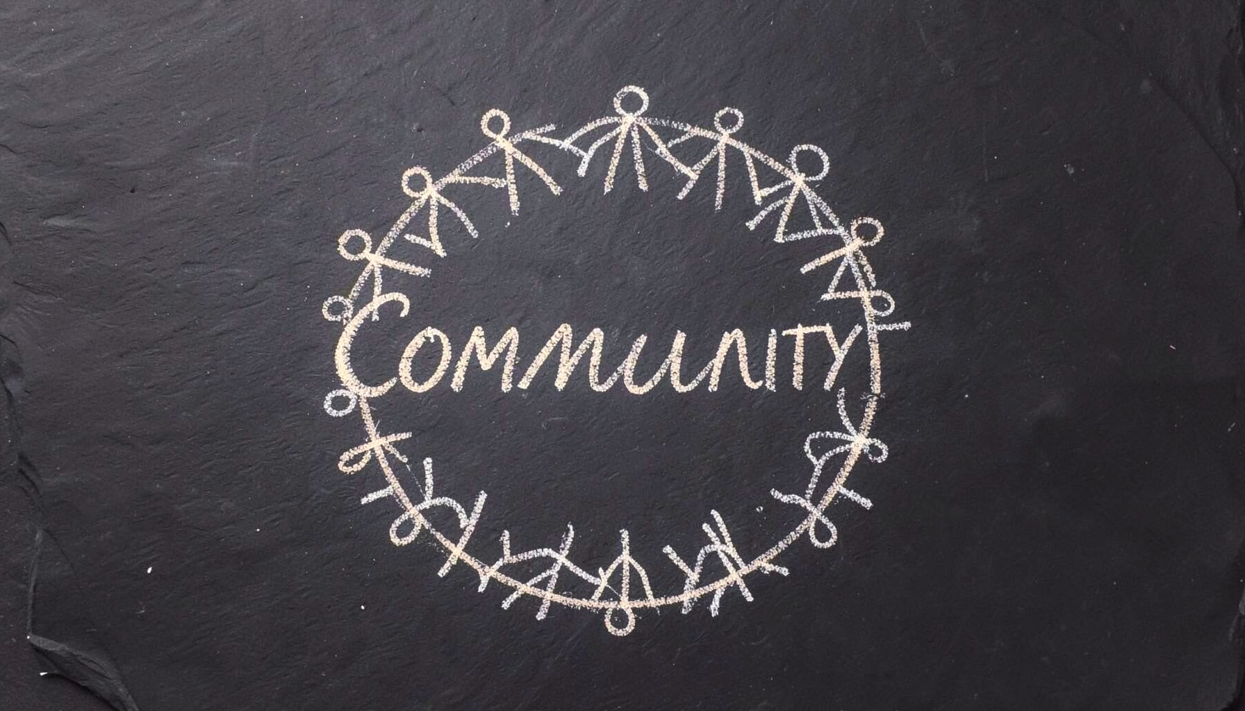 A chalk drawing on a dark slate board showing a circle of connected stick figures with the word COMMUNITY written in looping cursive chalk, warm white chalk lines on deep charcoal slate.