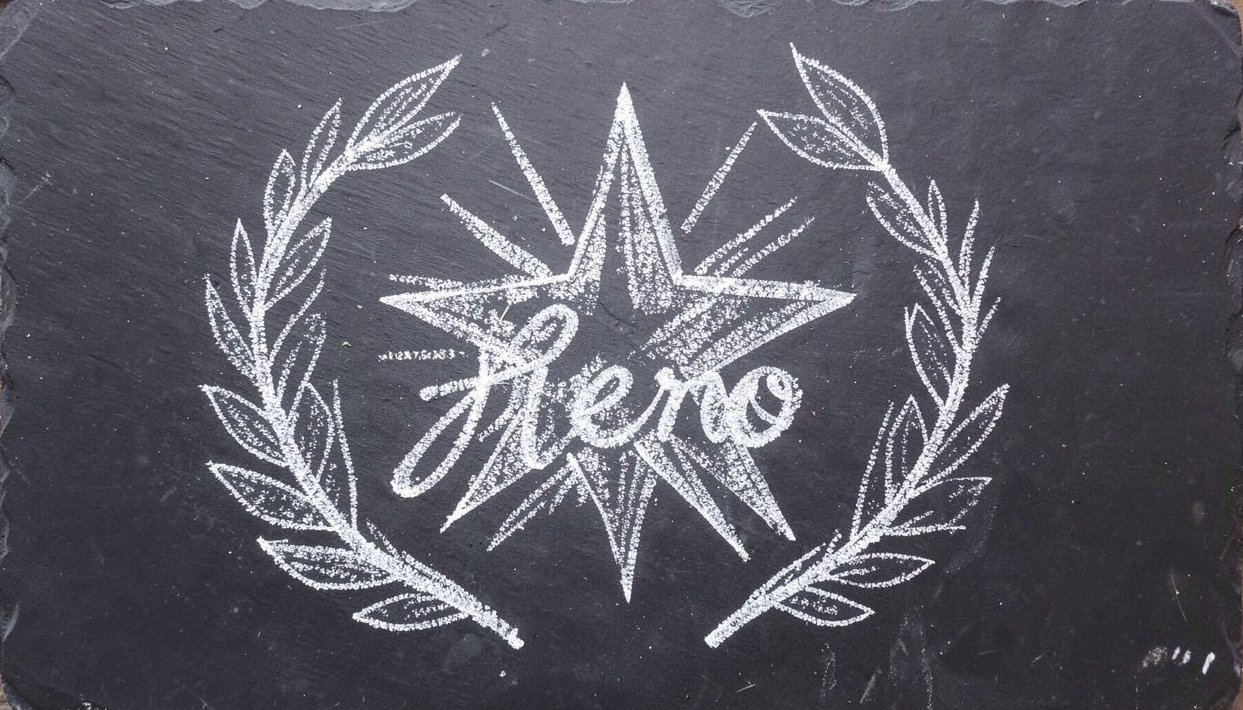 A chalk drawing on a dark slate board showing a large star with rays radiating outward, surrounded by hand-drawn laurel branches and the word 'Hero' sketched in cursive chalk lettering.